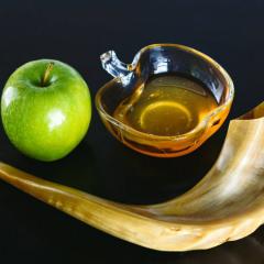 Whole green Apple sitting next to a glass bowl with golden honey. Both are sitting with the curve of a shofar or ram’s horn used to call Jews to worship on Rosh Hashanah