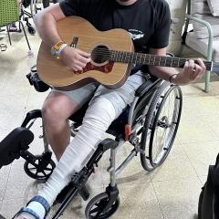 Young man sitting in a wheelchair holding a guitar on his bandaged leg. You can't see his face for privacy reasons